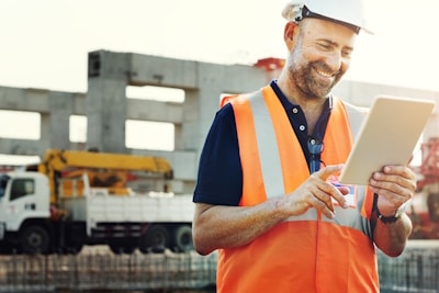 A construction worker in a helmet and orange vest uses a tablet at a building site, accessing and viewing resources.